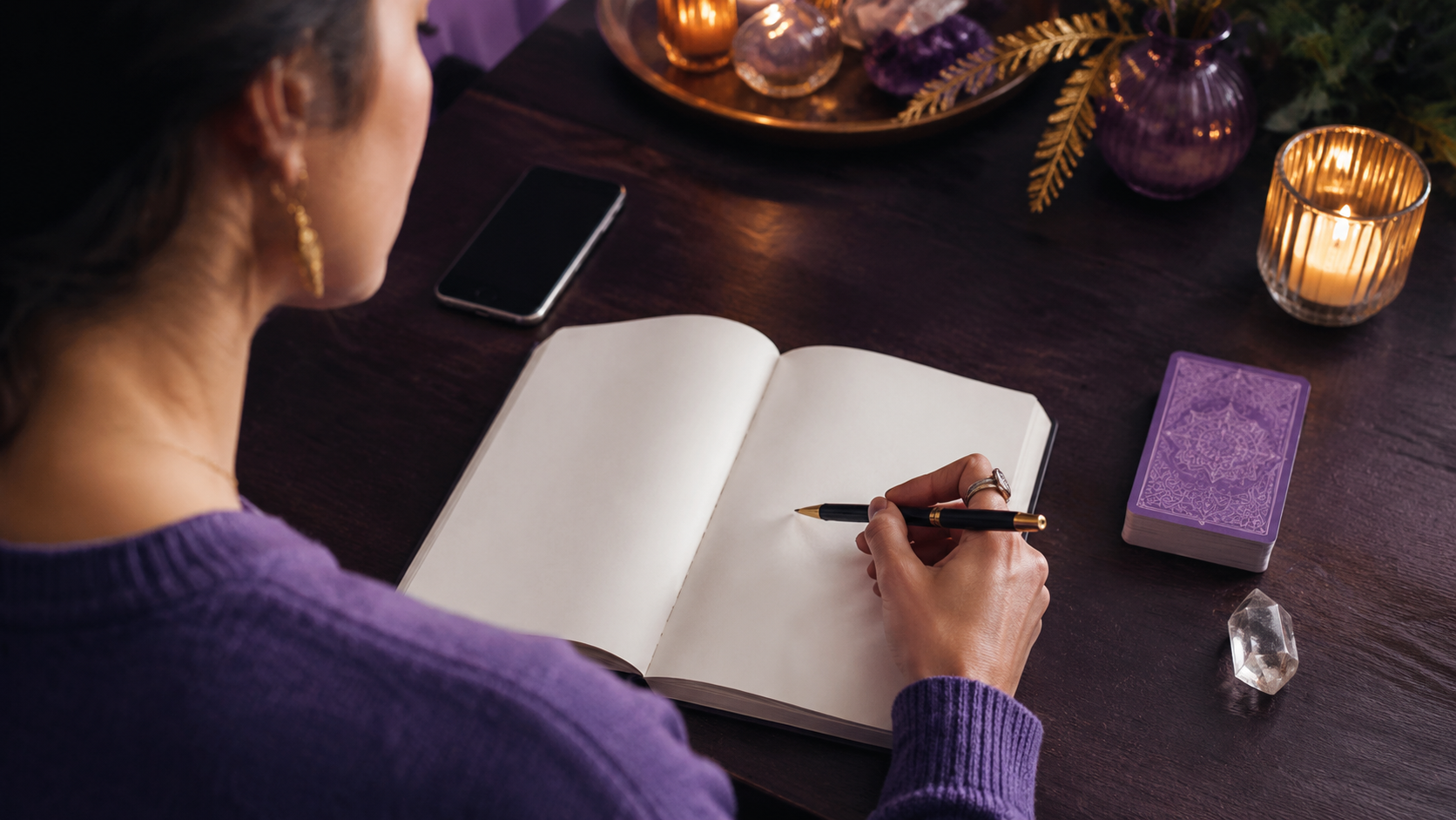 Woman writing psychic reading questions in a blank journal beside a phone, tarot deck and clear quartz